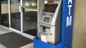Ein Geldautomat mit blauem Sockel und weißer 'ATM'-Beschriftung, vor einem Gebäude mit Glastüren gelegen.