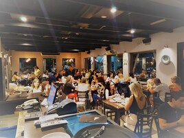 Un restaurante concurrido con varias mesas y sillas, un mostrador de recepción con teléfono y ordenador, y amplias ventanas que dejan entrar luz natural.