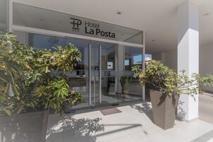 La entrada del Hotel La Posta, con fachada de vidrio, plantas en macetas grandes y vista despejada del interior a través de las puertas de cristal.