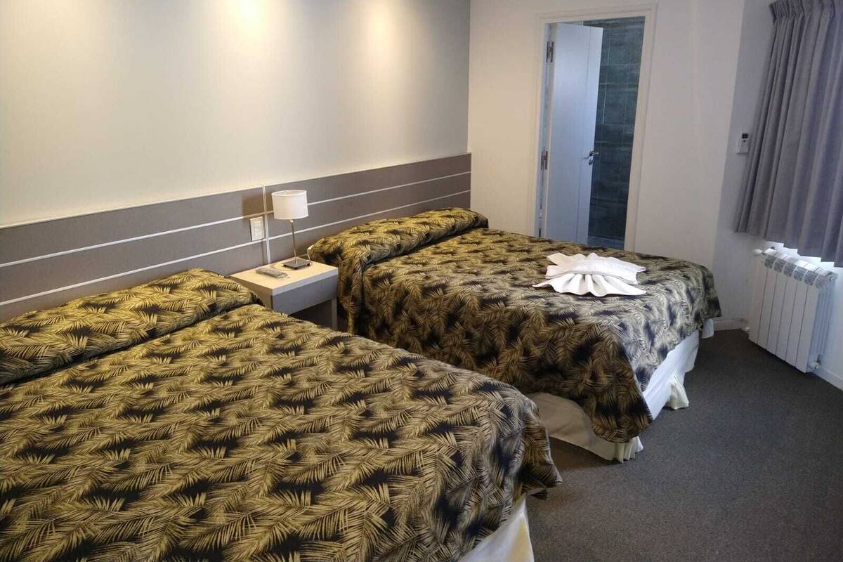 Camera d'albergo con due letti singoli, un comodino con lampada, un termosifone e una porta che conduce al bagno.