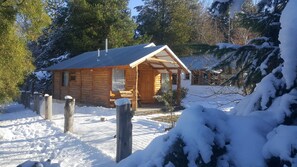 A wooden cabin with a porch, surrounded by snow-covered trees and a fenced yard. - Cabanas La Cautiva (Esquel)