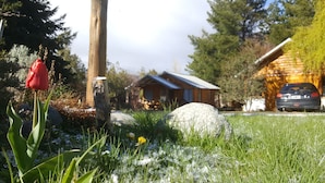 Un chalet rustique avec un toit en pente, entouré d’un jardin où l’on peut voir des tulipes rouges, de gros rochers et une voiture garée au loin.