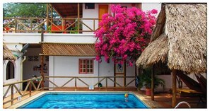Ein Swimmingpool mit einer strohgedeckten Struktur, ein Balkon mit Geländer und eine leuchtend rosa Bougainvillea-Pflanze.