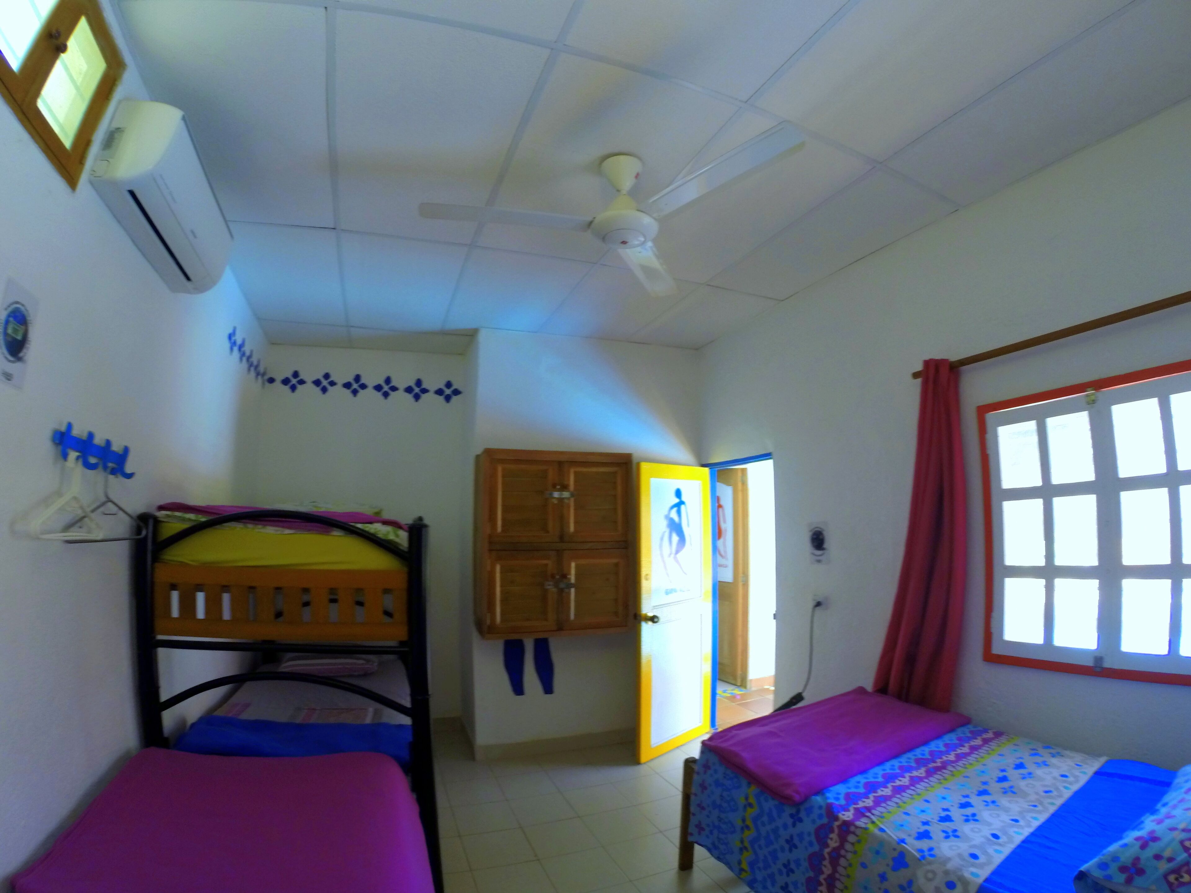Quarto com beliches, ventilador de teto, ar condicionado, janela com cortinas e porta que dá para outro cômodo.