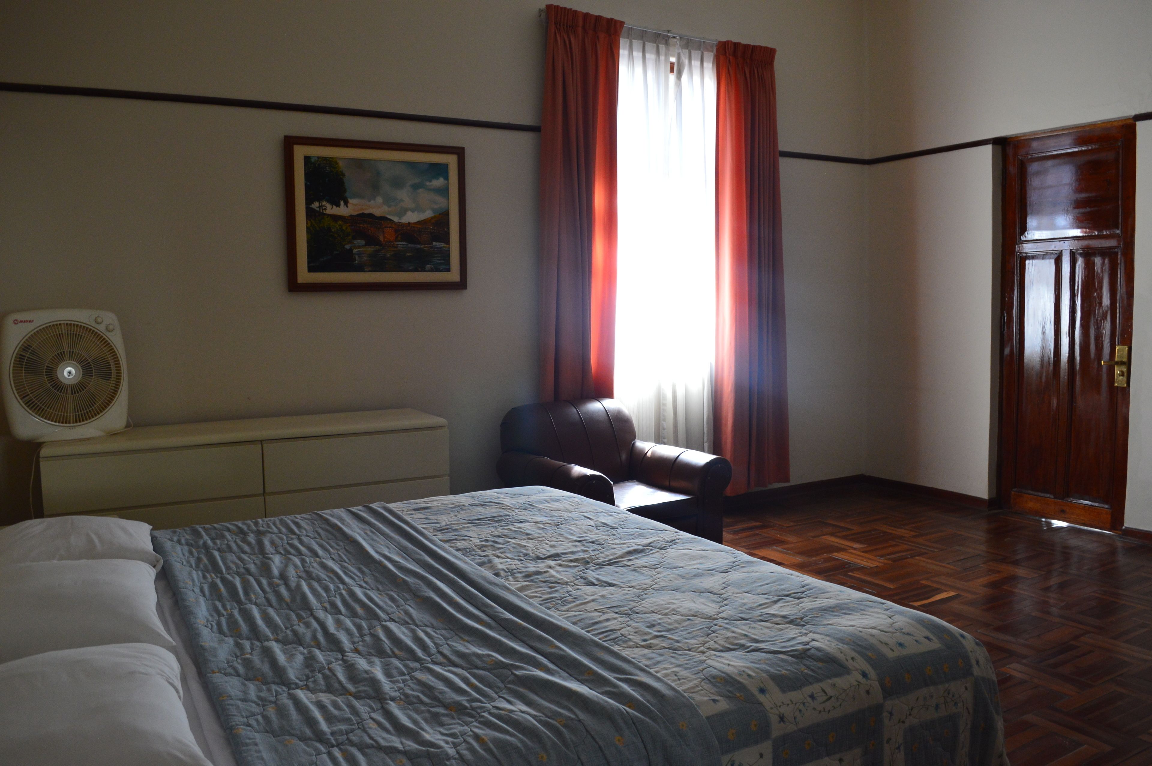 A bedroom with a bed, a chair, a fan, a painting, and a door.