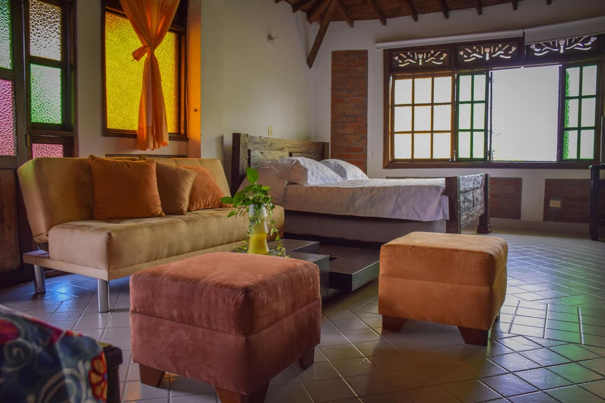 A room with a sofa, ottoman, and bed, a window with a view, and a brick wall.