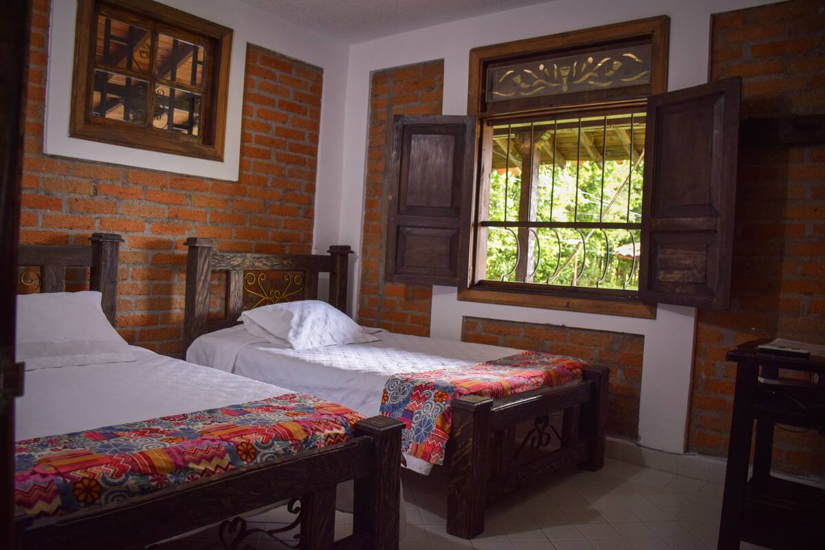 A room with two wooden beds, a brick wall, and a window with a view of greenery.