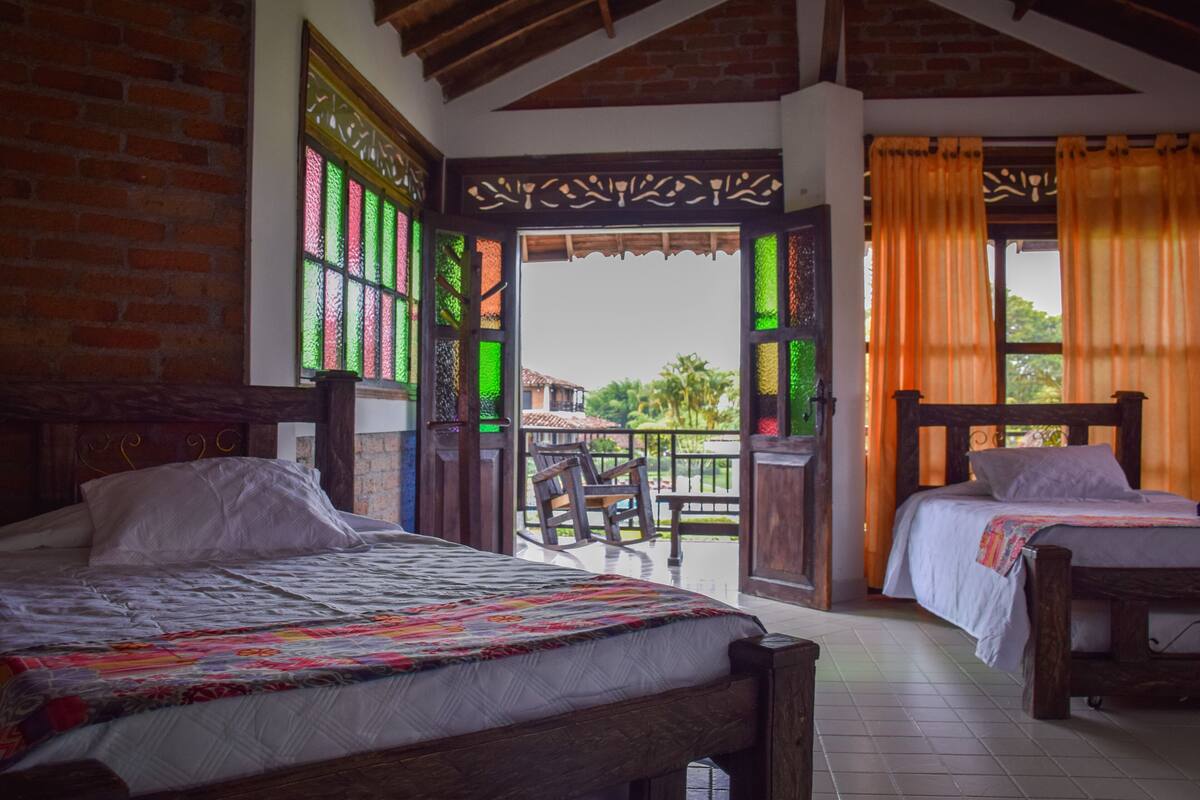 A room with two wooden beds, a balcony with a view, and a decorative window with green and red patterns.