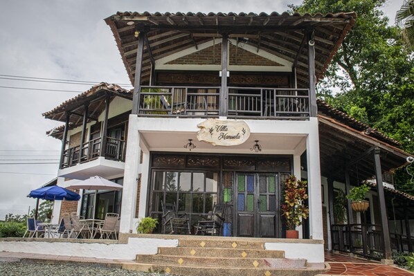 A two-story building with a balcony, a sign reading "Villa Manuel," outdoor seating with umbrellas, and a potted plant. - Villa Manuela - Finca Hotel Casa Nostra (Quimbaya)