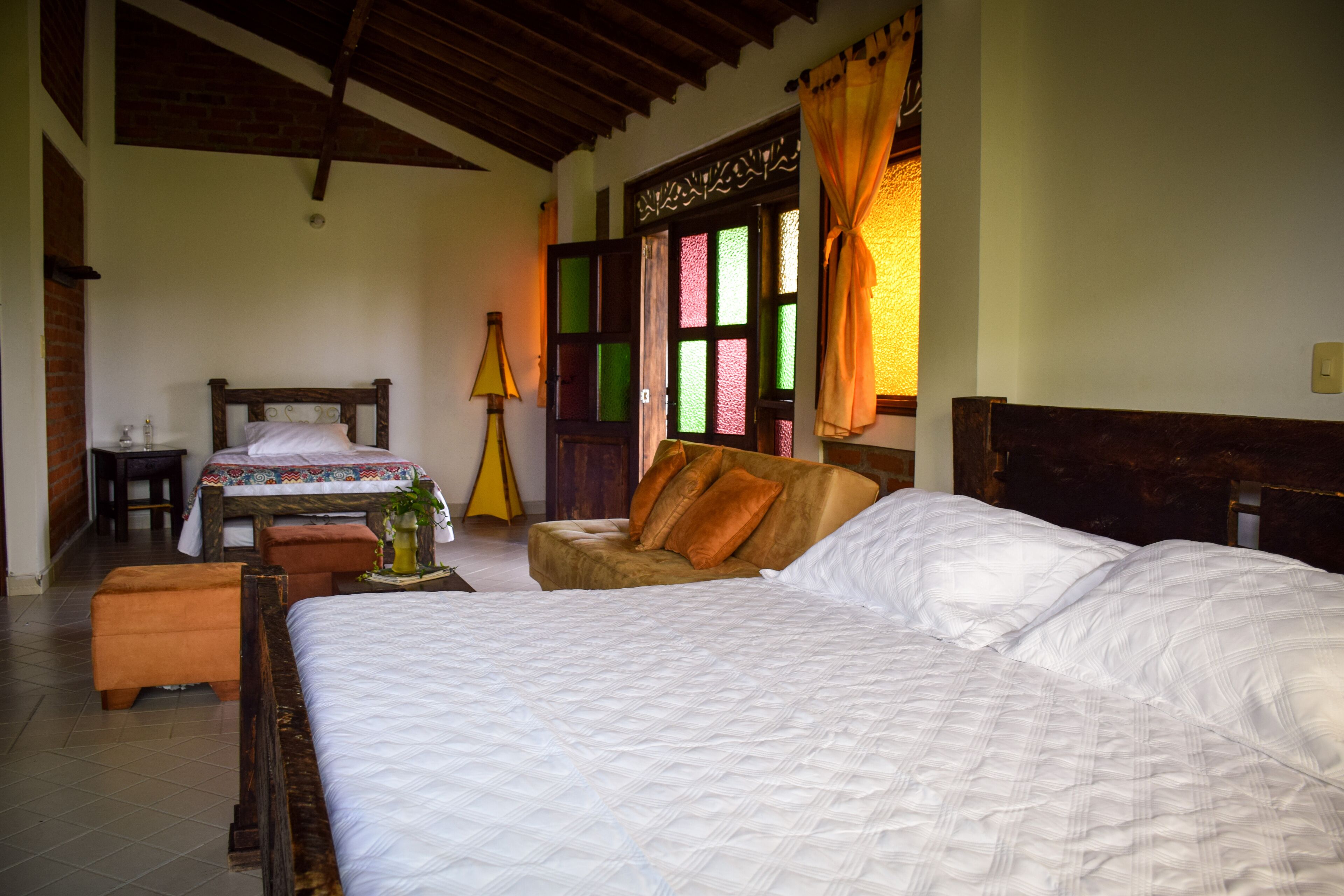 A bedroom with a large bed, a wooden headboard, a small table with a lamp, and a window with colorful panes.