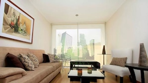 Uma sala de estar moderna com um sofá, uma mesa de centro e uma cadeira. Há uma janela grande com vista para prédios.