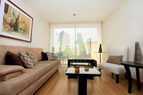 Un salon moderne avec un canapé, une table basse et un fauteuil. Une grande fenêtre offre une vue sur les immeubles.