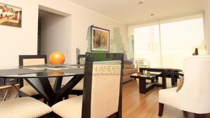 Uma sala de estar moderna com uma mesa de jantar de vidro, cadeiras acolchoadas, um sofá branco e uma obra de arte com vista para a cidade na parede.