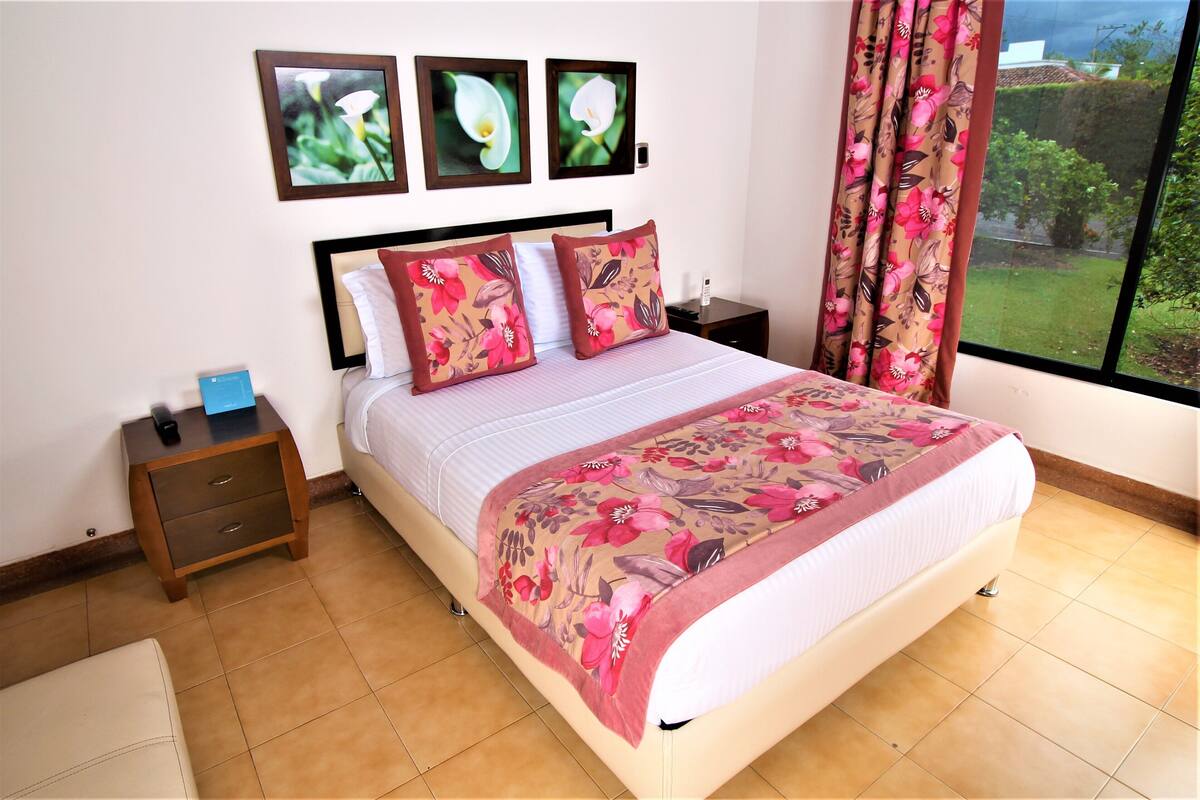 Een slaapkamer met een bed, nachtkastjes, een raam met gordijnen en ingelijste foto’s aan de muur.