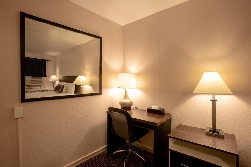 Una habitación de hotel con un escritorio, dos lámparas, una silla, un espejo y una mesita de noche.