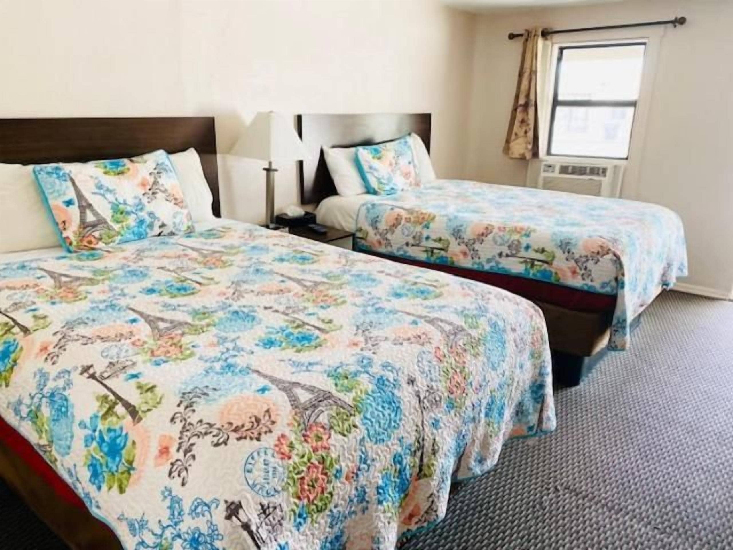 A hotel room with two beds, each with floral bedding, a headboard, bedside tables, and a window with curtains.