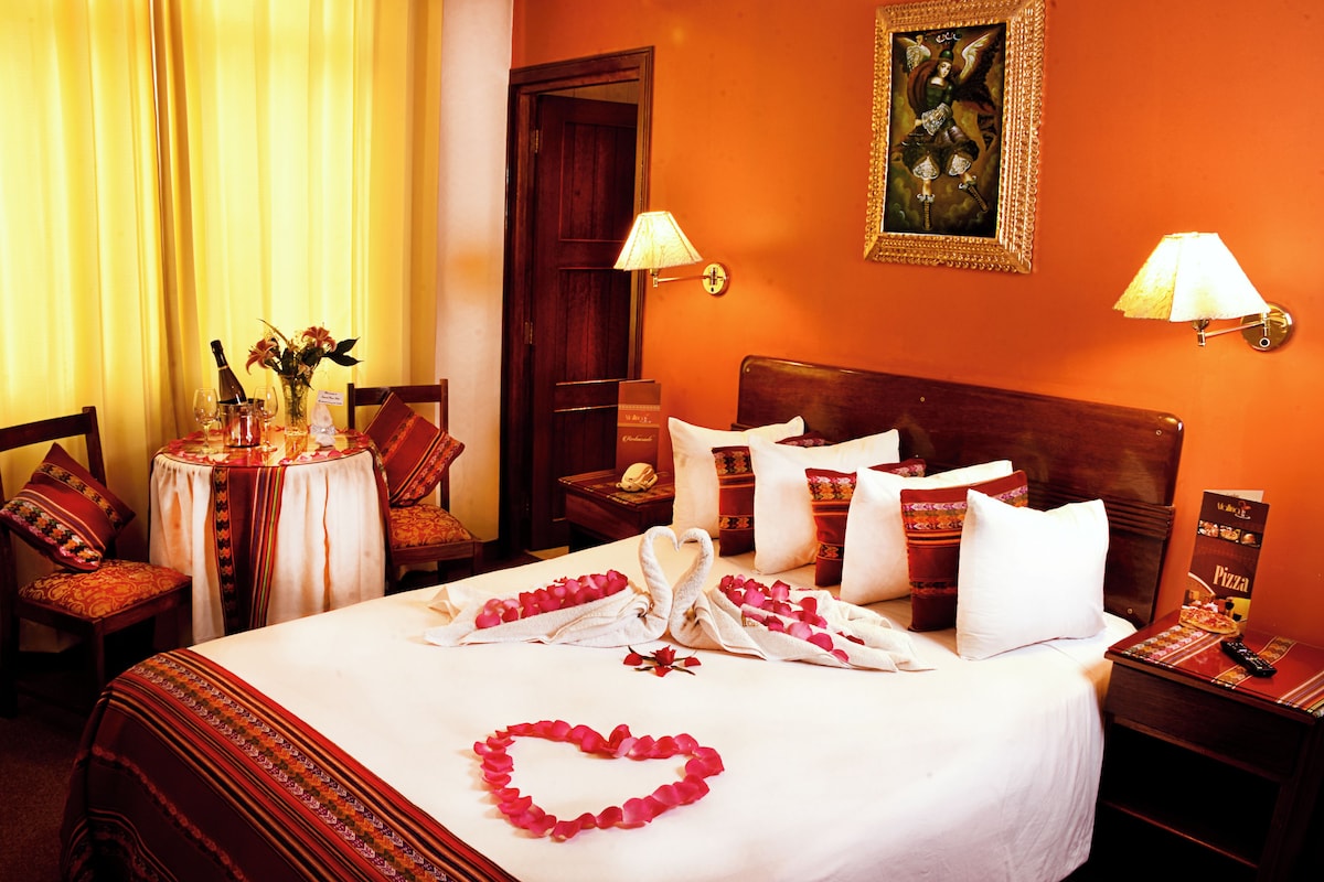 Una habitación de hotel con una cama bien hecha, una mesita redonda con un florero y una silla con cojín.