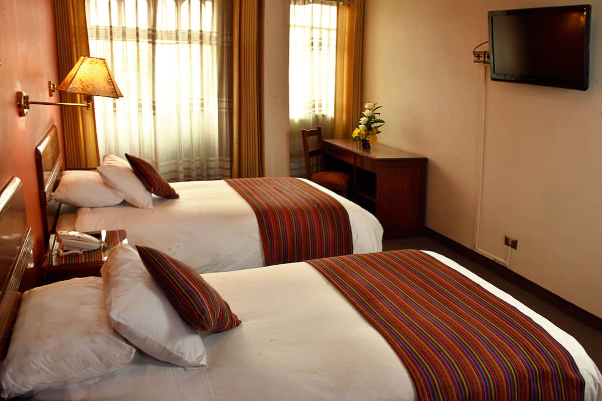 A hotel room with two beds, a desk, a chair, a TV, and a lamp.