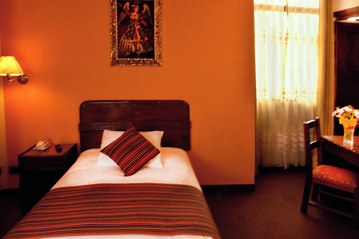 Habitación de hotel con cama, mesita de noche con lámpara, una silla y una ventana con cortinas.