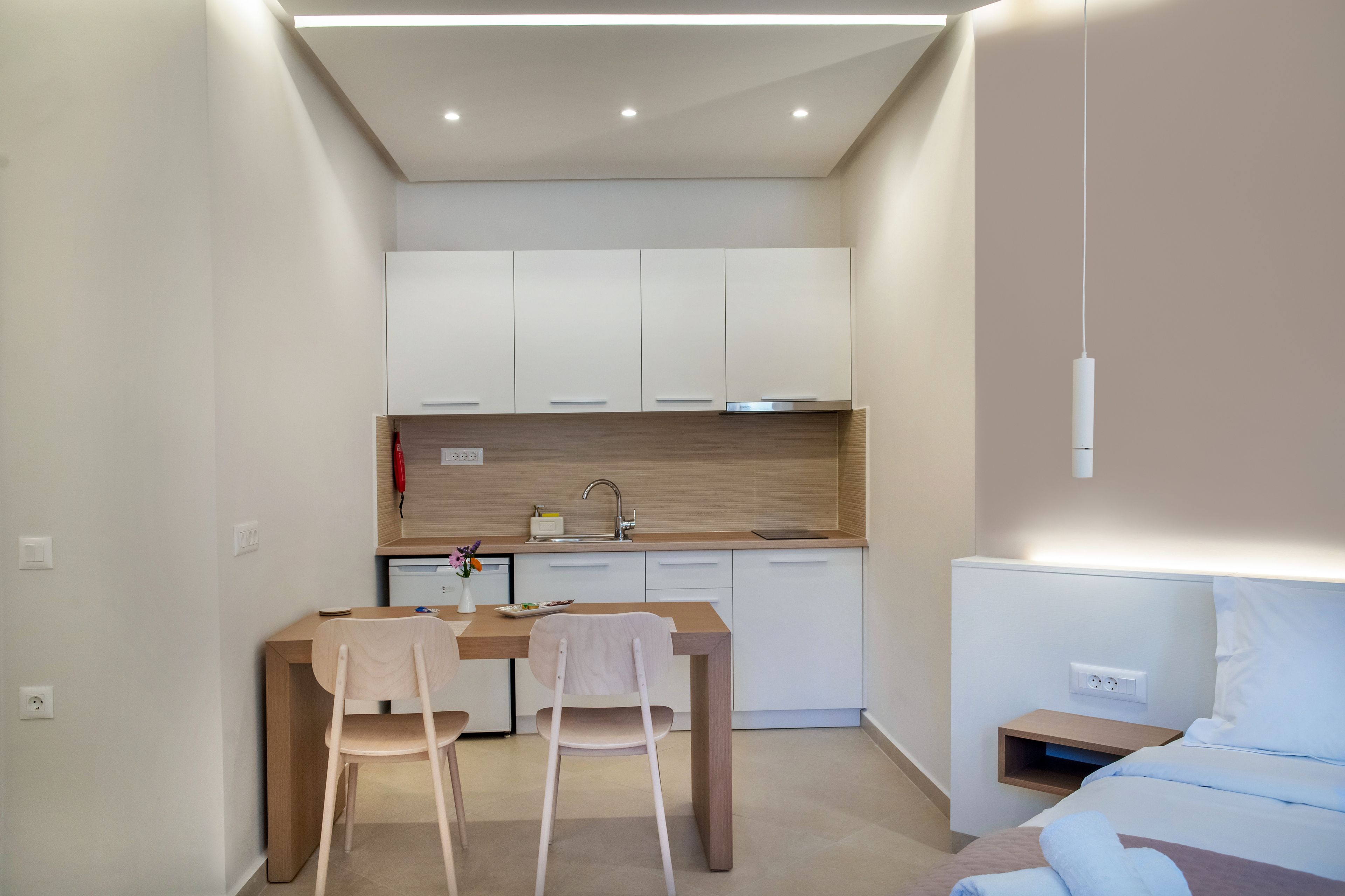 A compact hotel room with a kitchenette, a dining area with two chairs, a bed, and a bedside table.