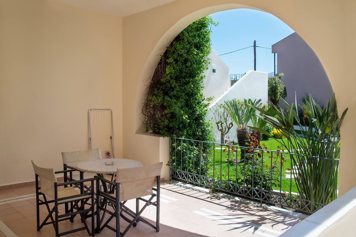 A balcony with a table and chairs, a view of a garden, and a white wall with greenery.