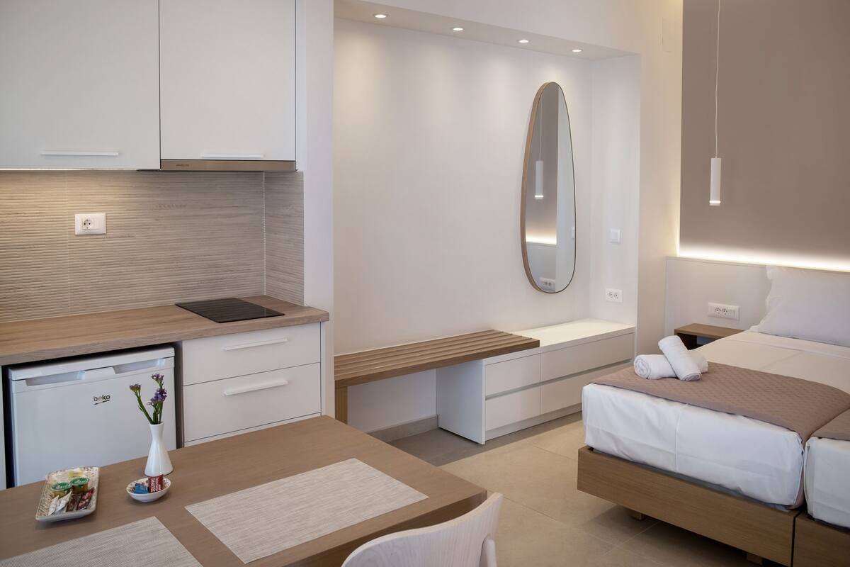 A modern hotel room with a kitchenette, a bed with a headboard, a bedside table, and a mirror.