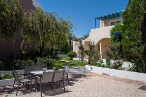 An outdoor patio with a table and chairs, surrounded by greenery and a clear sky.