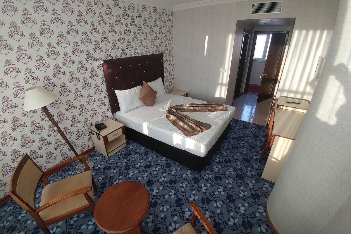 Una habitación de hotel con cama, mesita de noche, silla, una pequeña mesa y papel tapiz estampado.