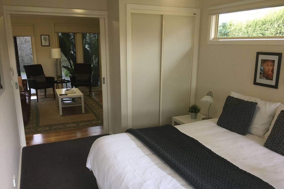 Un dormitorio con cama, mesita de noche, lámpara, una pequeña mesa con silla y vistas a un balcón.