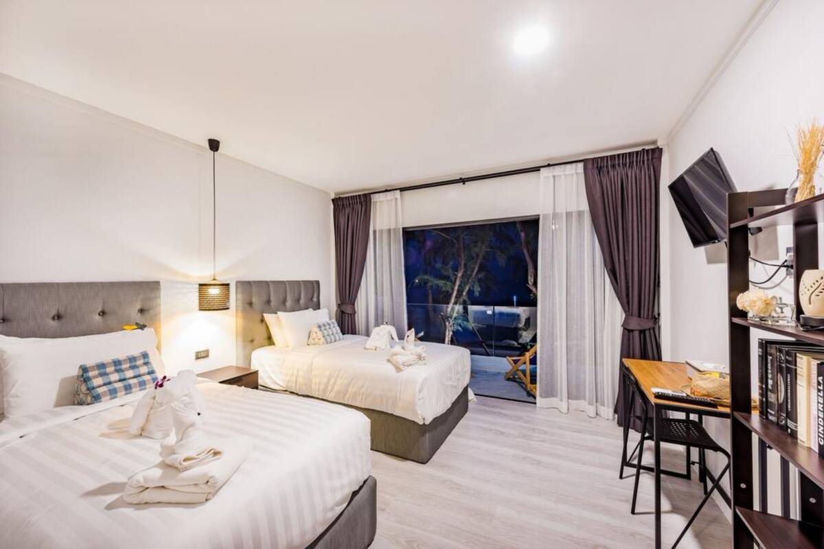 Quarto de hotel com duas camas, uma escrivaninha, uma cadeira, uma televisão e uma varanda com cortinas.