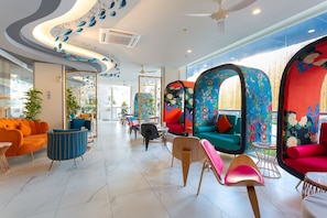 Un vestíbulo de hotel moderno con áreas de descanso coloridas, un ventilador de techo y un amplio ventanal con vistas al exterior.