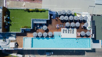 An aerial view of a rooftop pool area with a swimming pool, sun loungers, umbrellas, and a small lawn.