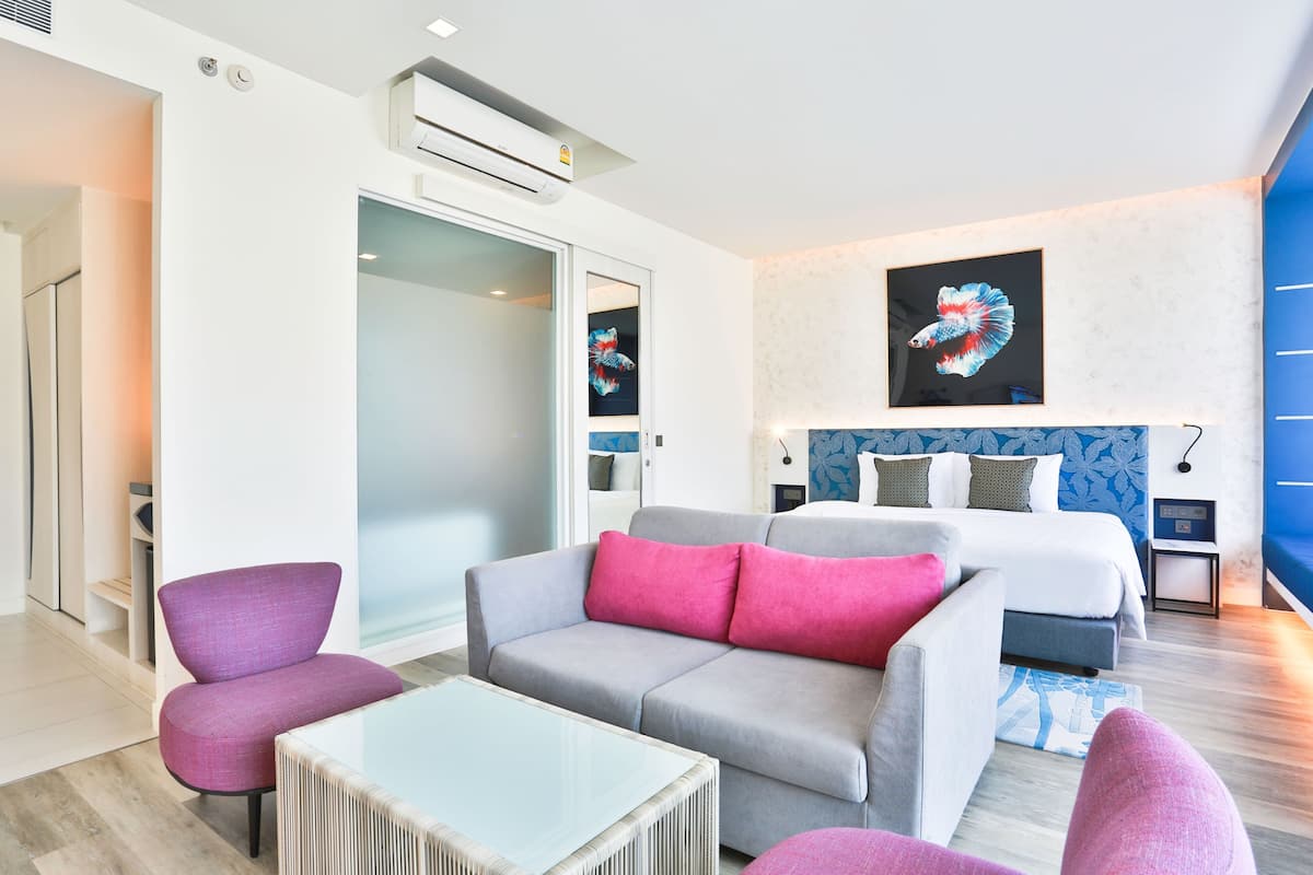 Una habitación de hotel moderna con un sofá gris, una pequeña mesa y una cama con cabecera azul. Hay una ventana grande y una puerta que conduce a un baño.