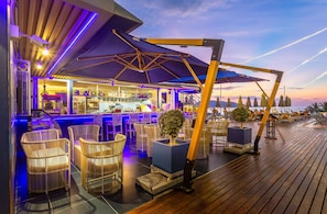 Zona de bar con sillas de mimbre y sombrillas, una barra con botellas y vista al mar.