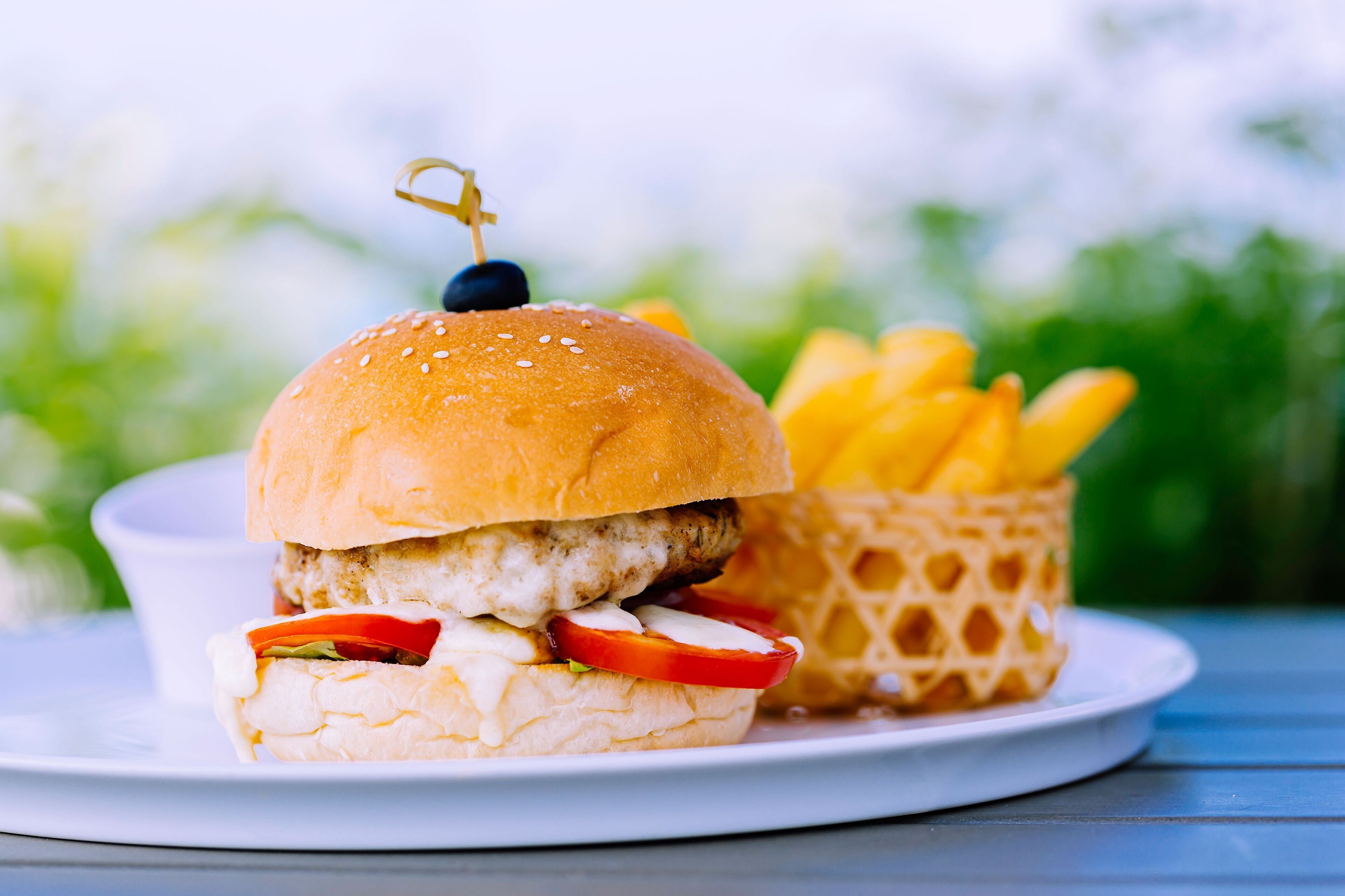 Ein Burger mit Sesambrötchen, einem Hähnchenbraten, Salat, Tomate und einem Stück Blauschimmelkäse, dazu eine Portion Pommes in einem Waffelbehälter.