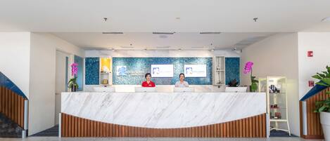 A reception desk with two staff members, a blue wall with a logo, and a potted plant.