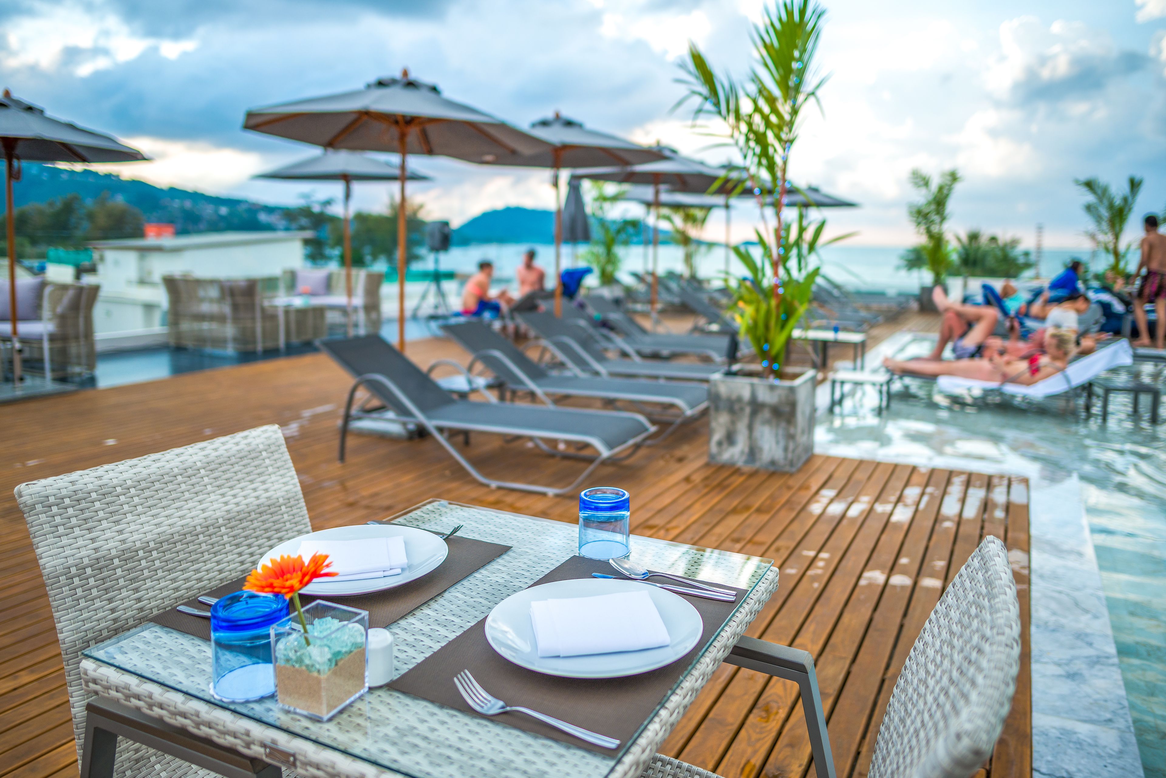 A dining table set with plates, cutlery, and a glass of water on a wooden deck overlooking a pool area with lounge chairs and umbrellas.