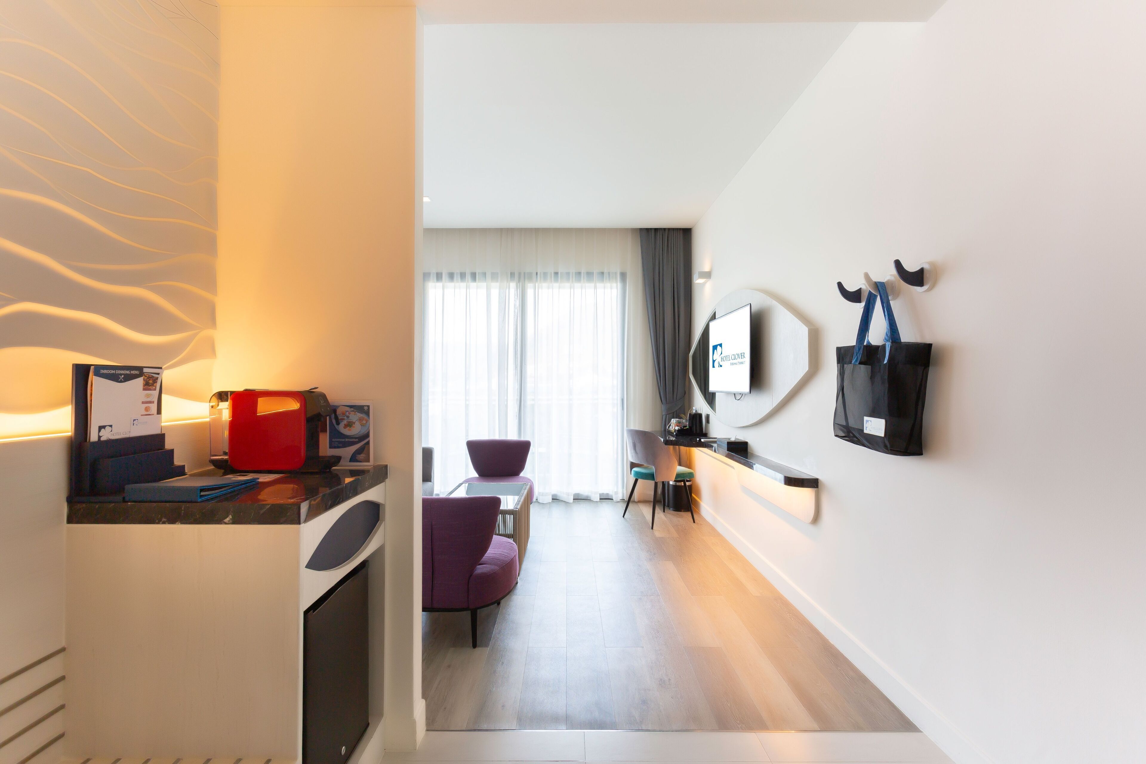 Une chambre d’hôtel moderne dotée d’un coin cuisine, d’une table à manger avec des chaises, d’un canapé, d’une grande fenêtre avec des rideaux et d’un téléviseur fixé au mur.