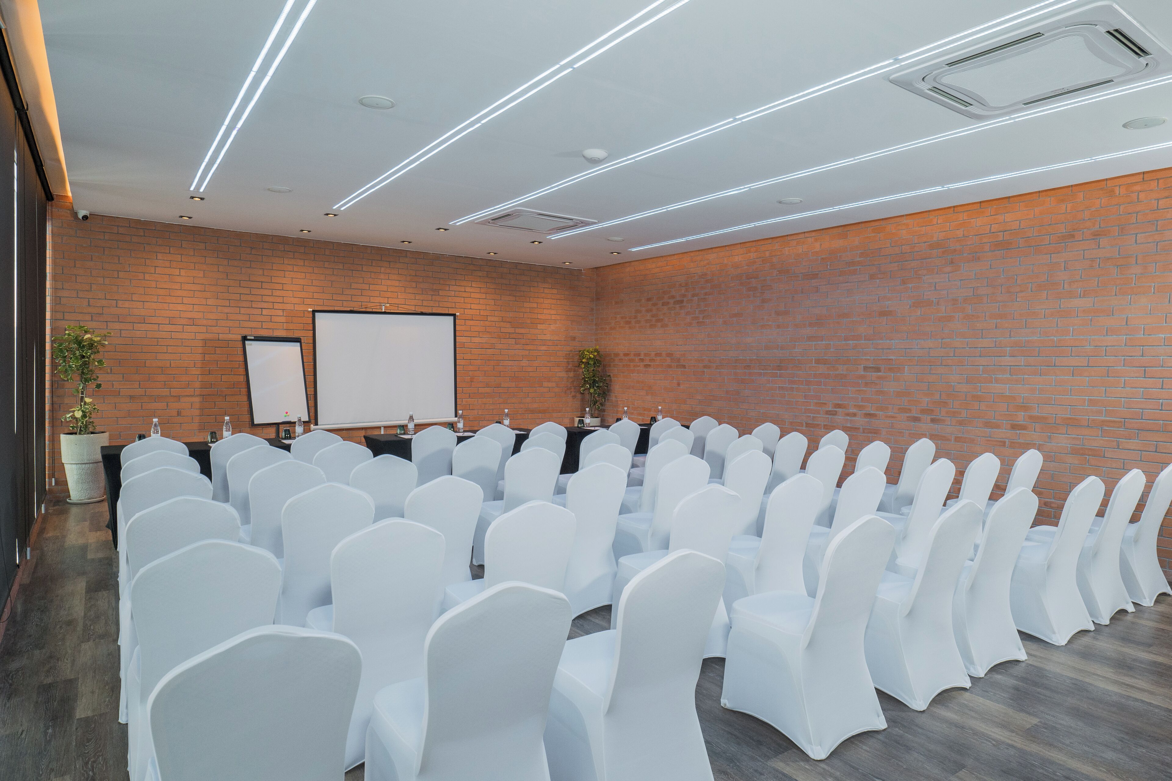 A conference room with rows of white chairs, a projector screen, and a brick wall.