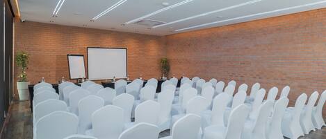 A conference room with rows of white chairs, a projector screen, and a brick wall.