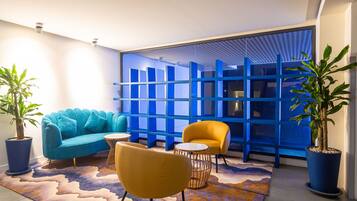 Un salon moderne avec un canapé bleu, un fauteuil jaune et une table basse ronde. On y trouve deux plantes en pot et un tapis à motifs sur le sol.