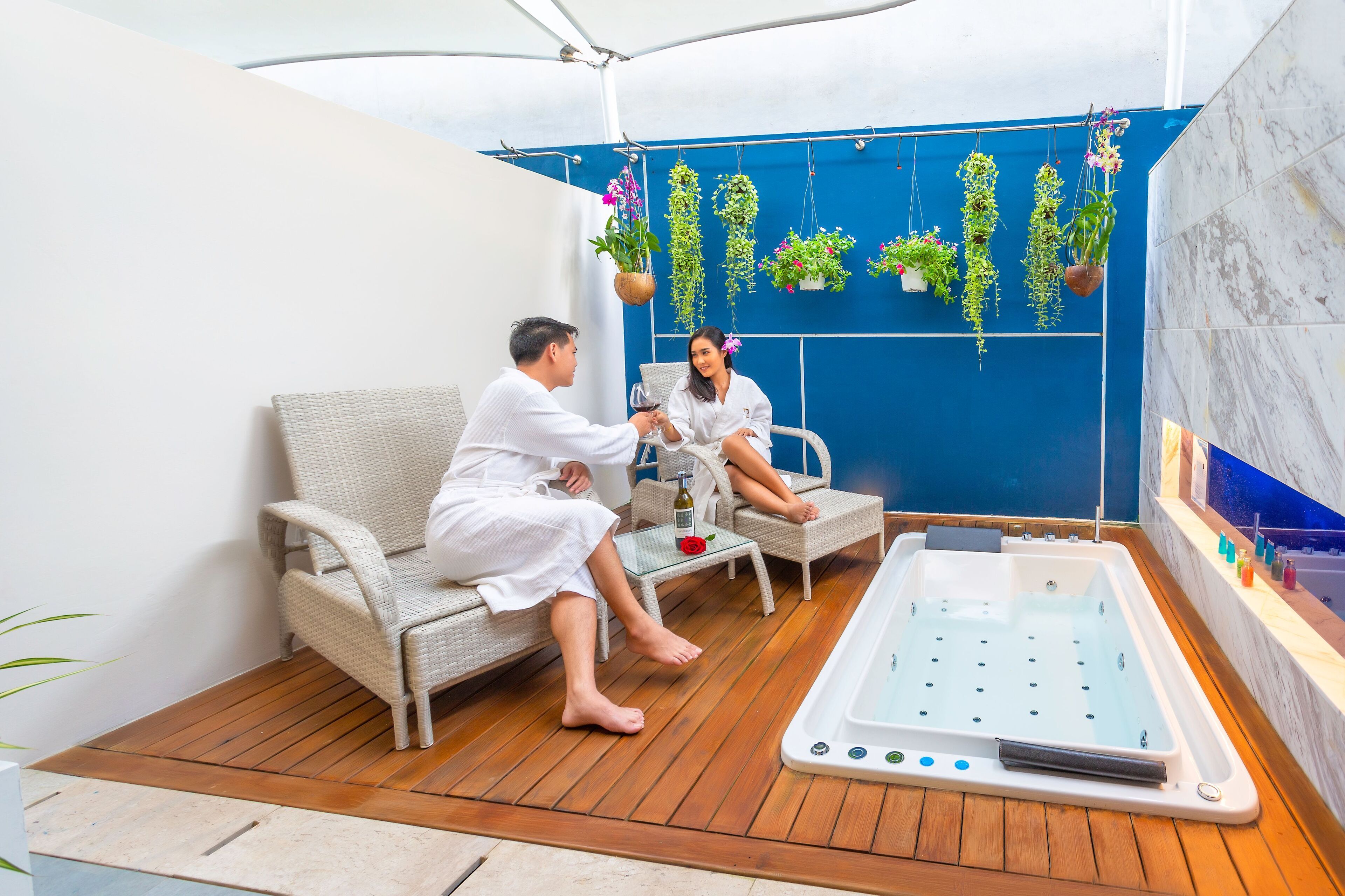 Un espace de bien-être avec deux personnes vêtues de robes blanches, un plancher en bois, une baignoire blanche et des plantes suspendues devant un mur bleu.