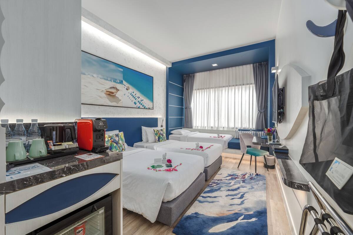 Habitación de hotel con dos camas, un escritorio con cafetera, televisión y un cuadro grande de una playa.