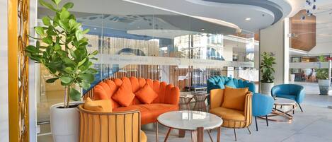 A modern lobby with a seating area featuring orange and blue chairs, a white coffee table, and a large potted plant.