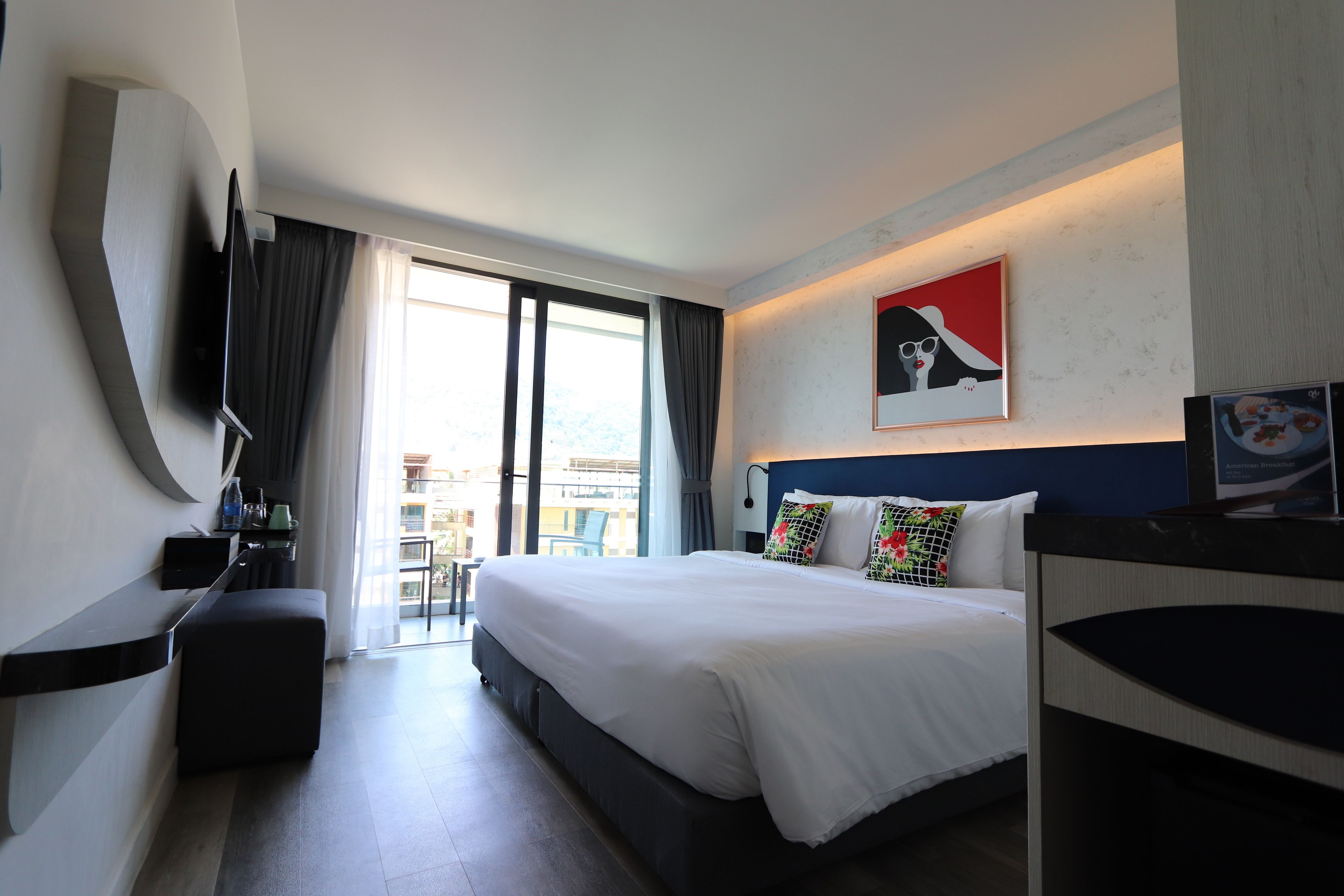 Habitación de hotel con una cama grande, un televisor de pantalla plana montado en la pared, un escritorio con una silla y vista a la ciudad por la ventana.