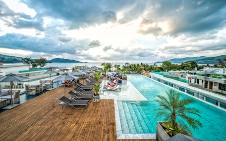 A rooftop pool area with lounge chairs, umbrellas, and a view of the sea and mountains.