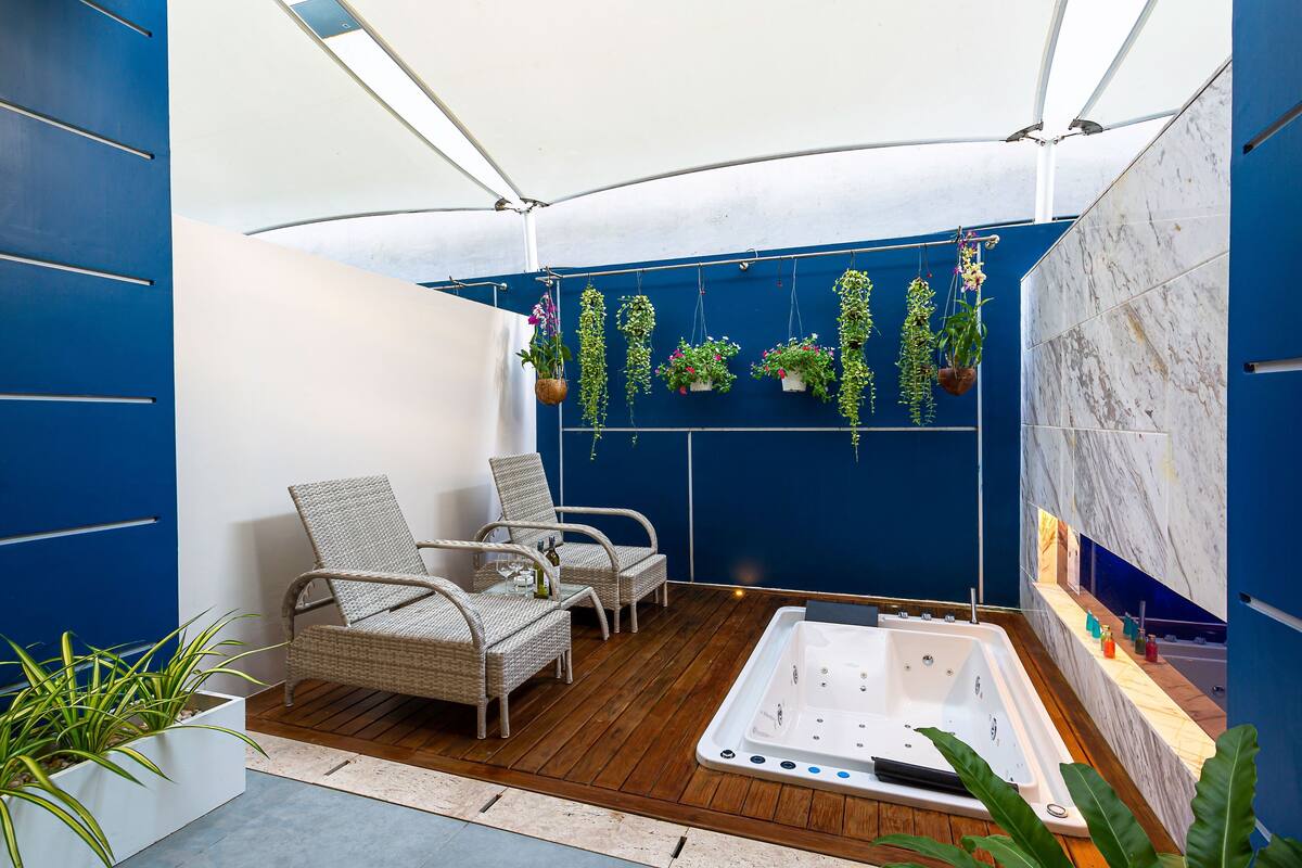 Un área de estar moderna al aire libre con sillas de mimbre, un jacuzzi y macetas colgantes frente a una pared azul.