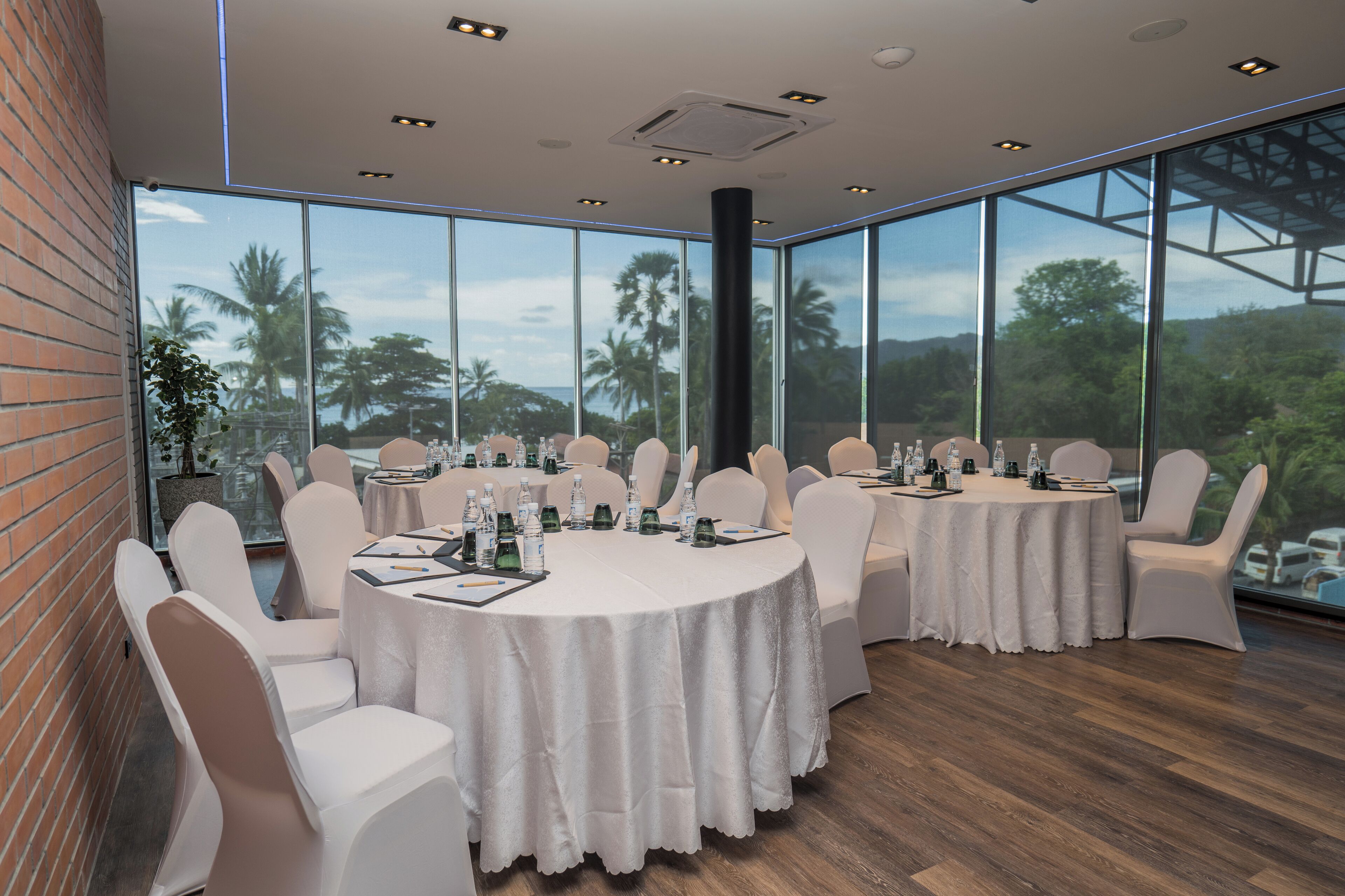 Un salón de conferencias con mesas redondas, sillas blancas y amplios ventanales que dan a vistas de palmeras y colinas.