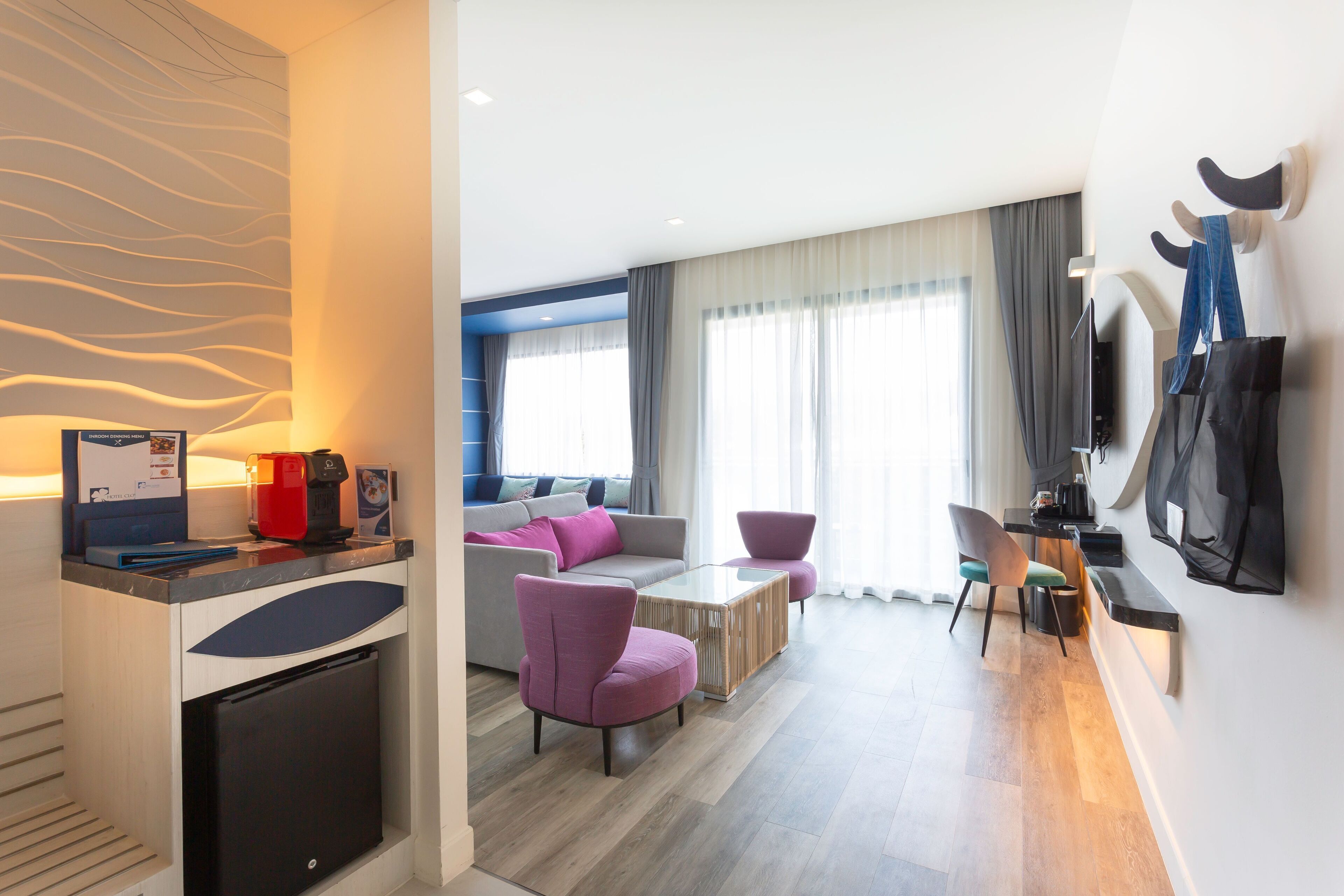 A modern hotel room with a sofa, chairs, a coffee machine, a TV, and a mini-fridge.