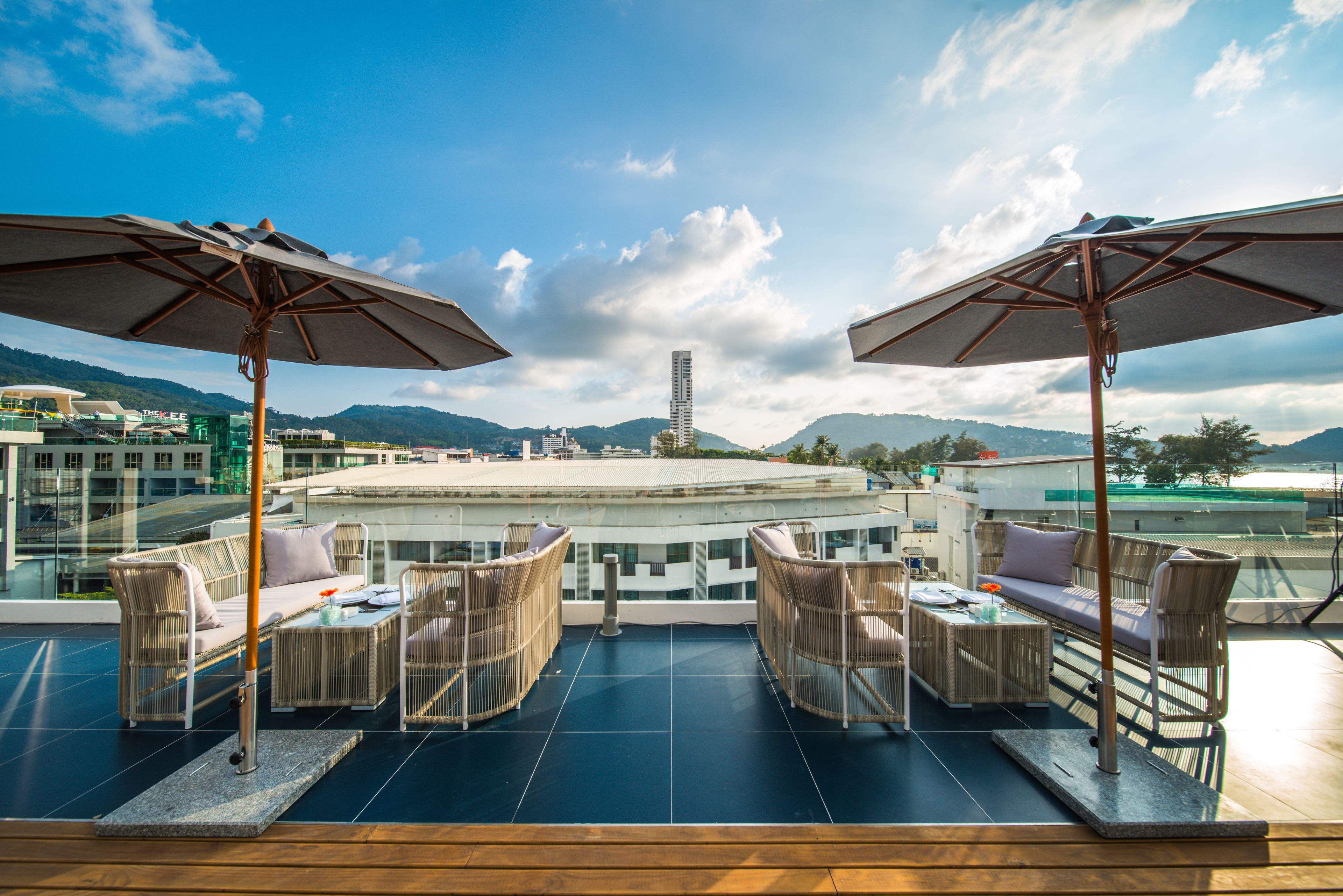 A rooftop terrace with wicker furniture, umbrellas, and a view of a cityscape and mountains.